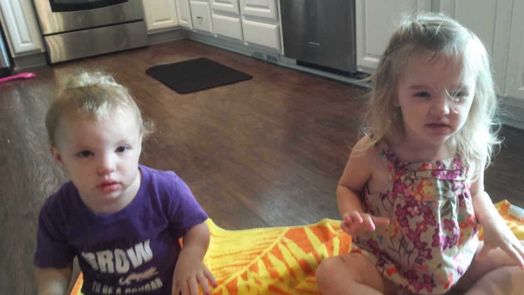 Two young children sitting on a colorful blanket in a kitchen.