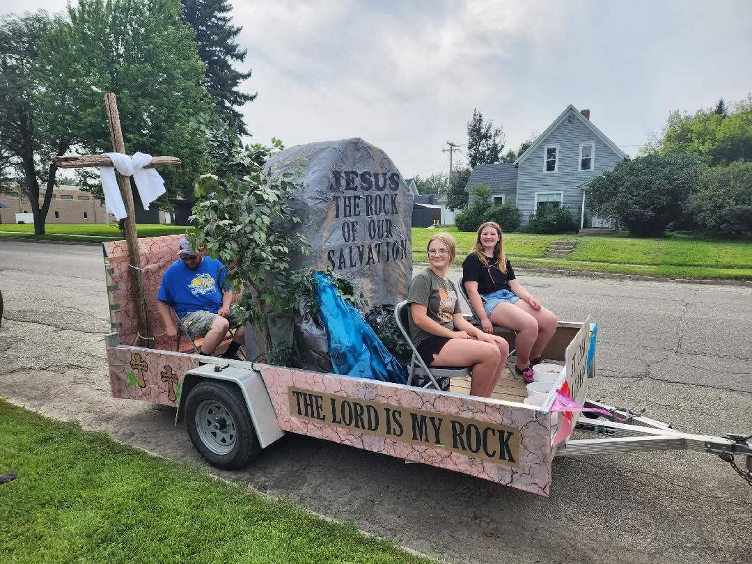 A decorated trailer featuring a large stone with the inscription 'Jesus, The Rock of Our Salvation' and a wooden cross, with three individuals sitting on it, positioned in a green outdoor setting.